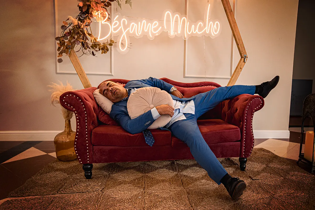 Novio tumbado en un sofá rojo bajo un neón de “Bésame mucho”